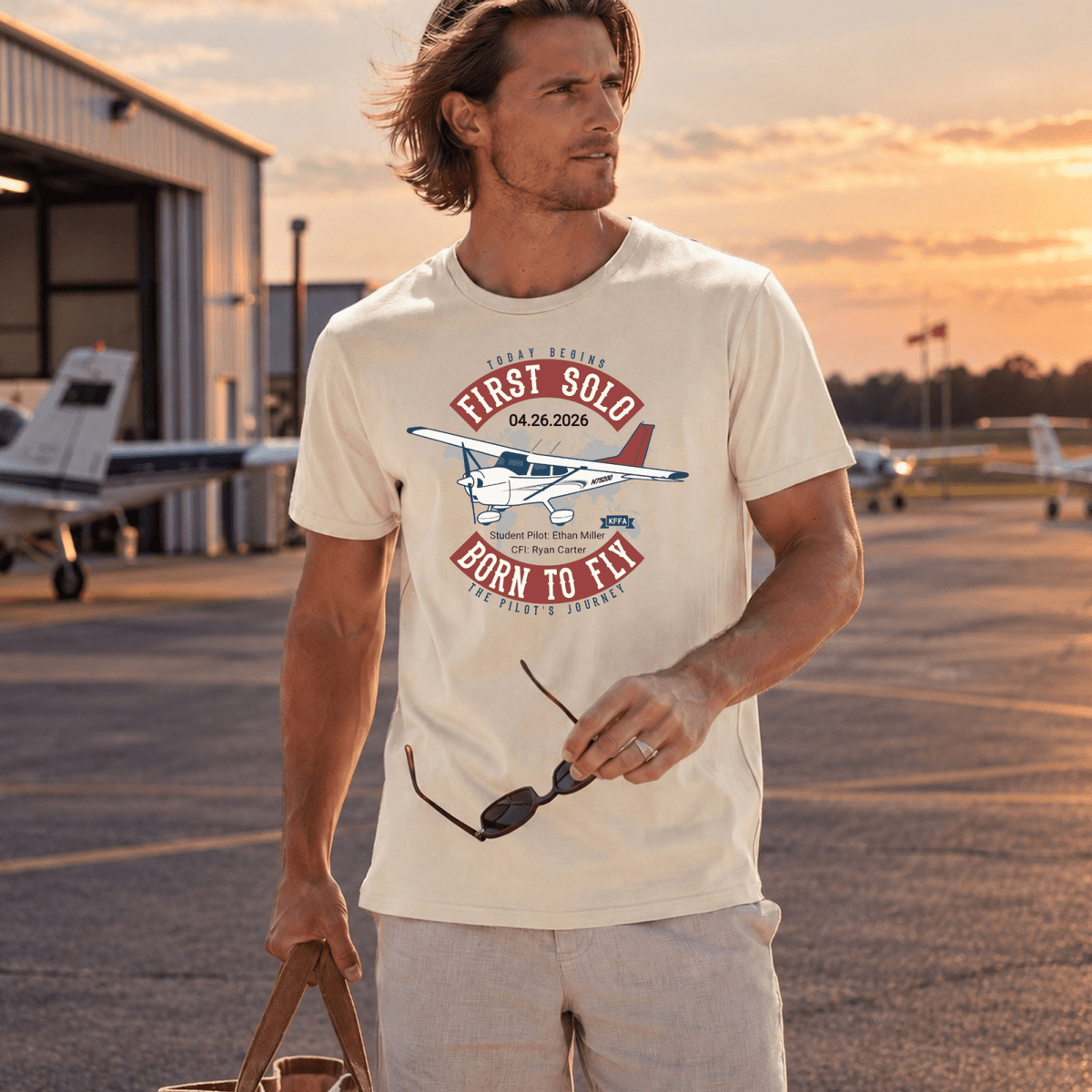 Man wearing a cream-colored First Solo Born to Fly T-shirt outside an aircraft hangar at sunset