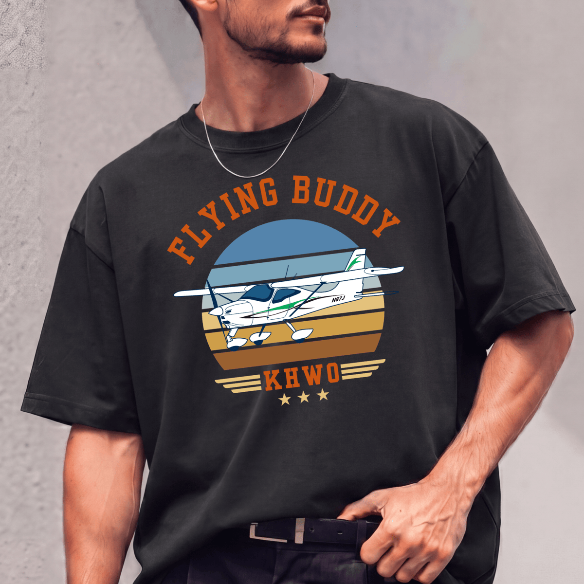 Man wearing a black Flying Buddy T-shirt with a white-and-green airplane against a gray wall