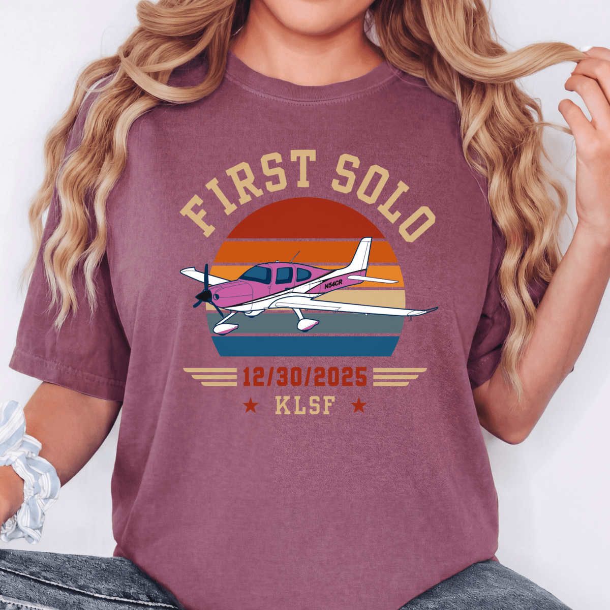Woman wearing a berry-colored First Solo T-shirt with a pink airplane and retro sunset design