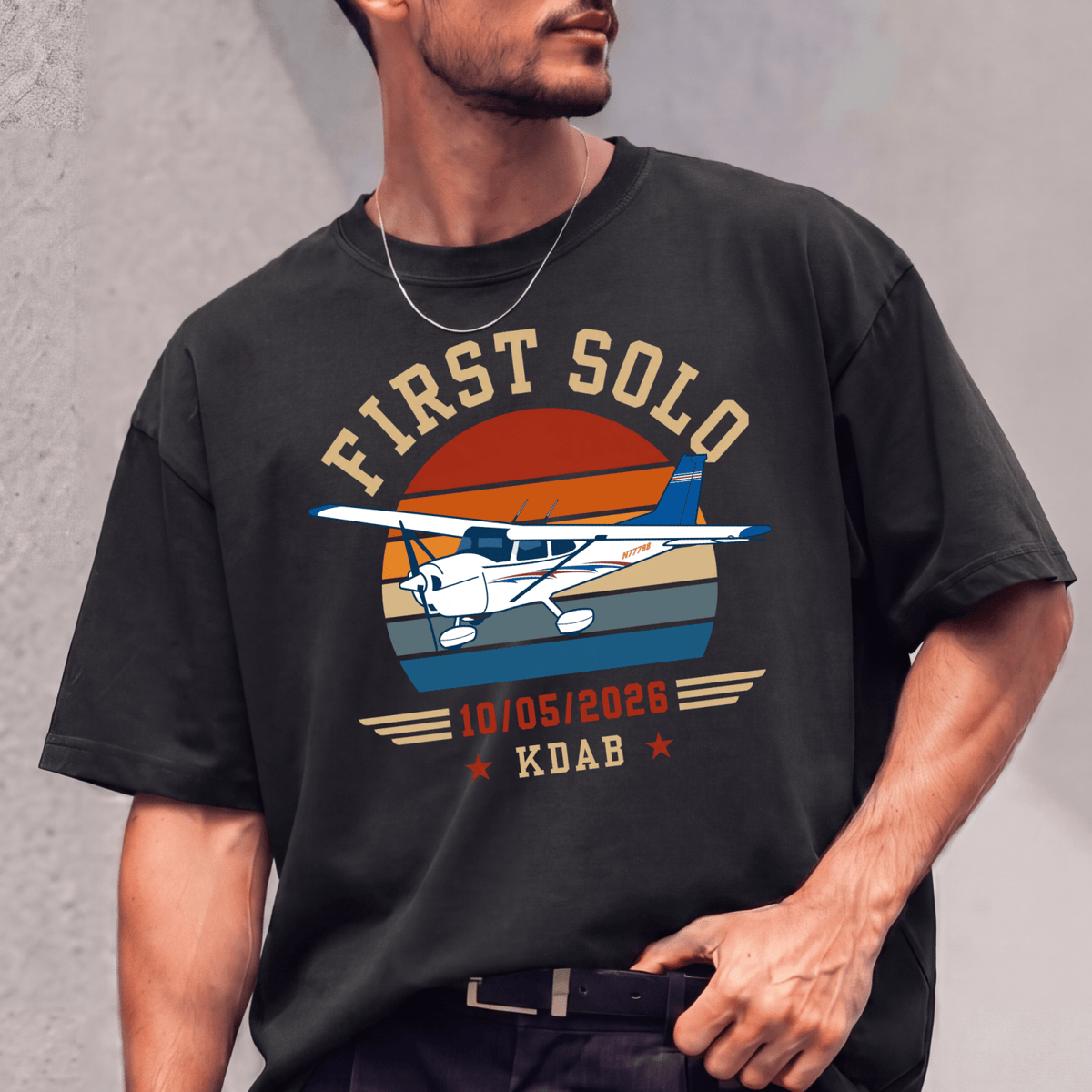 Man wearing a black First Solo T-shirt with a high-wing airplane and retro sunset graphic against a gray wall