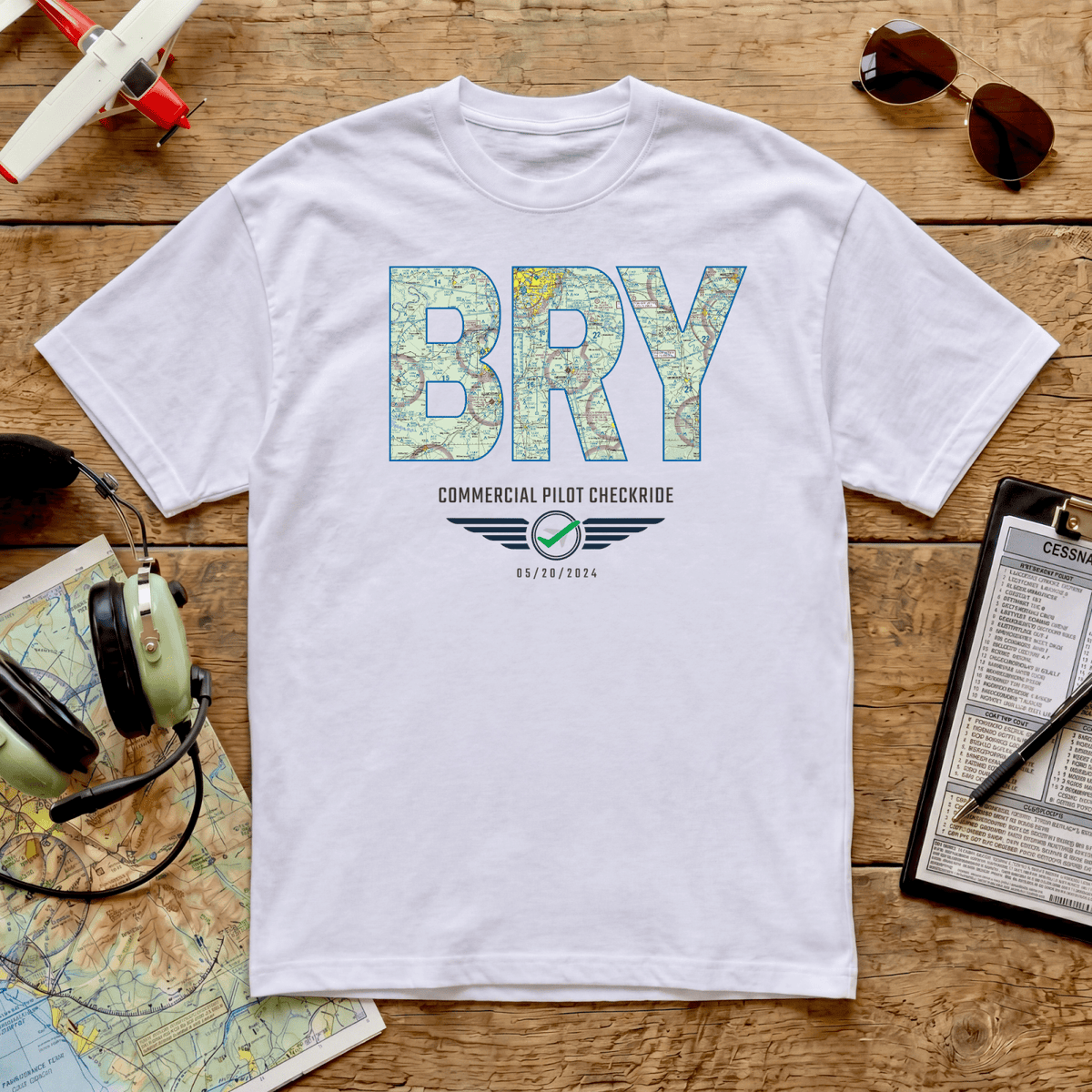 White BRY airport code VFR chart T-shirt celebrating a commercial pilot checkride, displayed on a wooden table