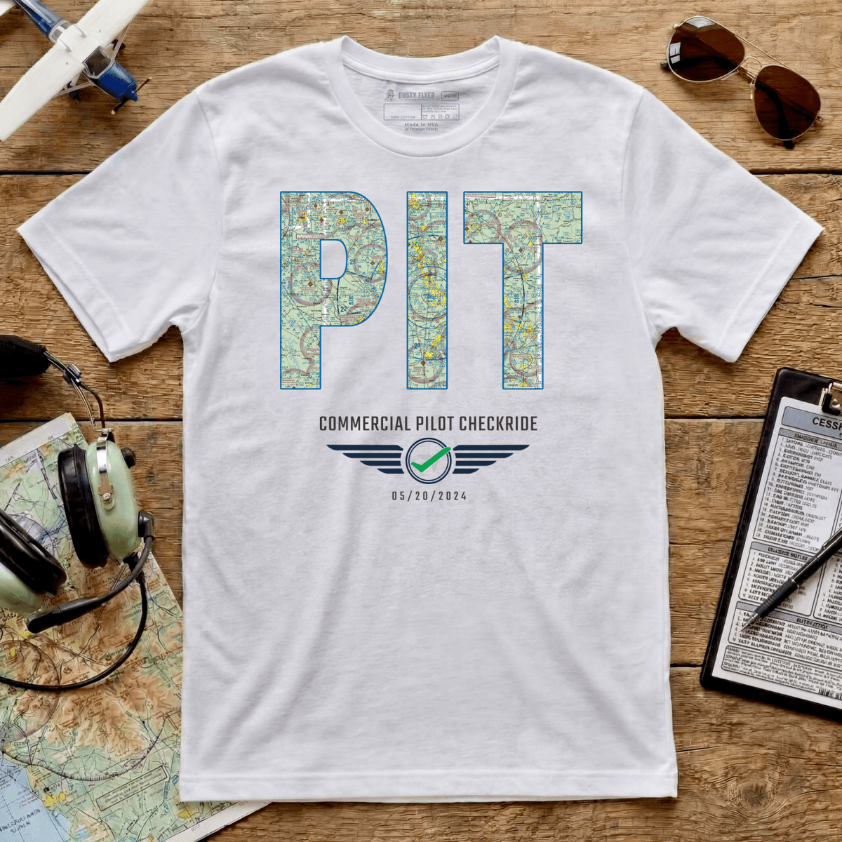 White PIT airport code VFR chart T-shirt with a commercial pilot checkride milestone beside aviation accessories